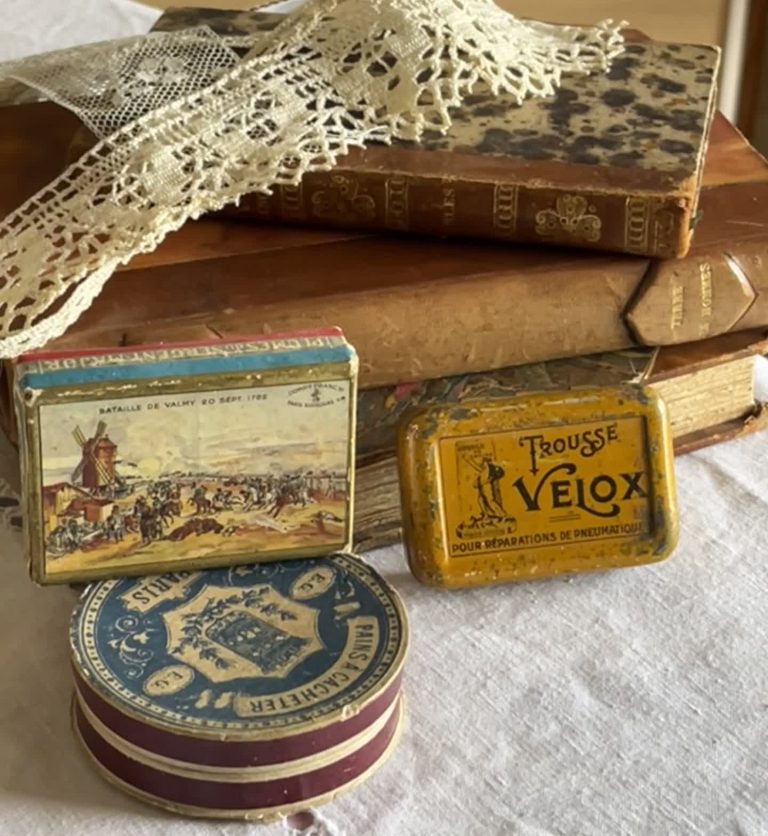 Livres anciens, boîtes métalliques vintage, et dentelle ancienne, posés sur un tissu clair