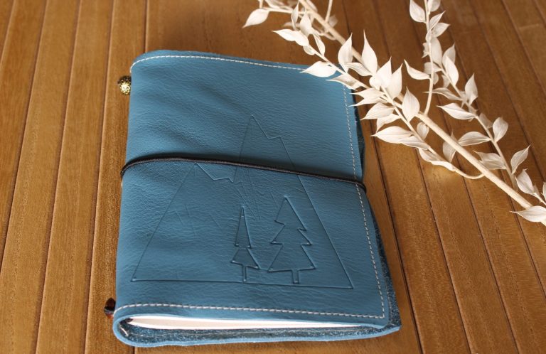 Carnet de randonnée en cuir bleu orné de sapins gaufrés, fermé par un élastique noir