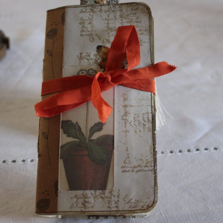 Carnet fermé noué d'un ruban rouge, orné d'un papillon et d'un motif de pot de plante, posé sur un tissu clair